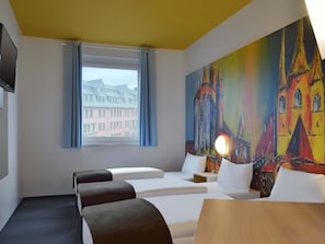 A hotel room with two beds, a wooden table, a painting of a colorful building, a window with a view of buildings, and a flat-screen TV on the wall.