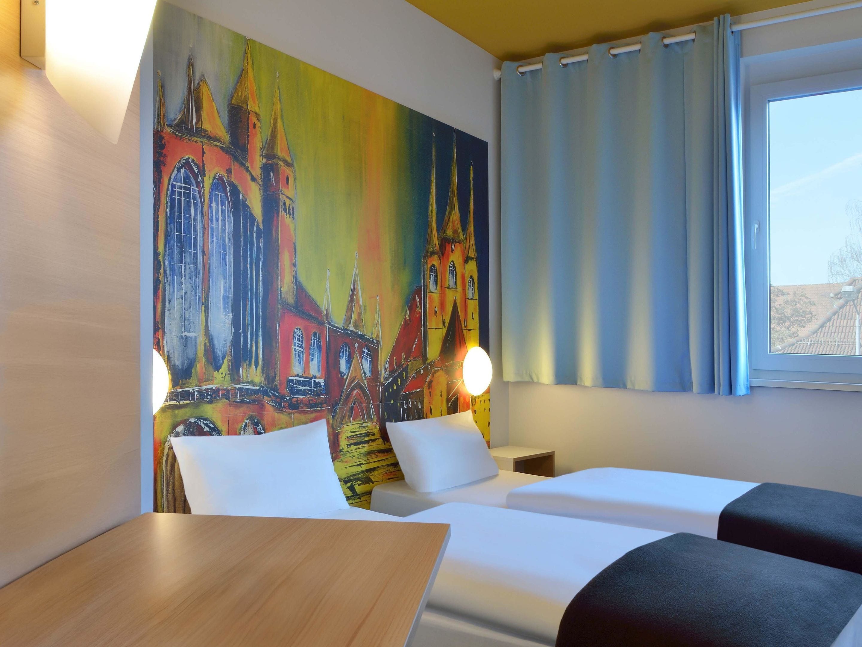 A hotel room with two beds, a painting of a church on the wall, a window with curtains, and a bedside table with a lamp.