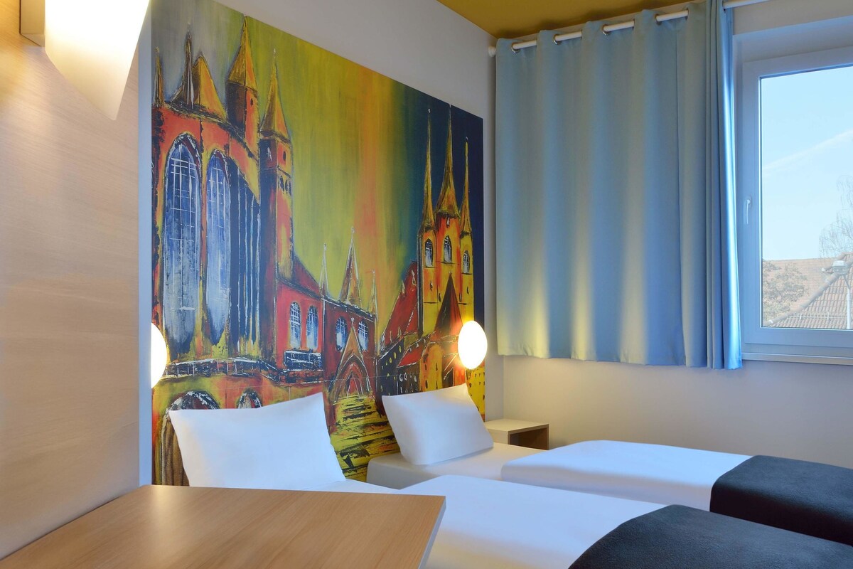 A hotel room with two beds, a painting of a church on the wall, a window with curtains, and a bedside table with a lamp.