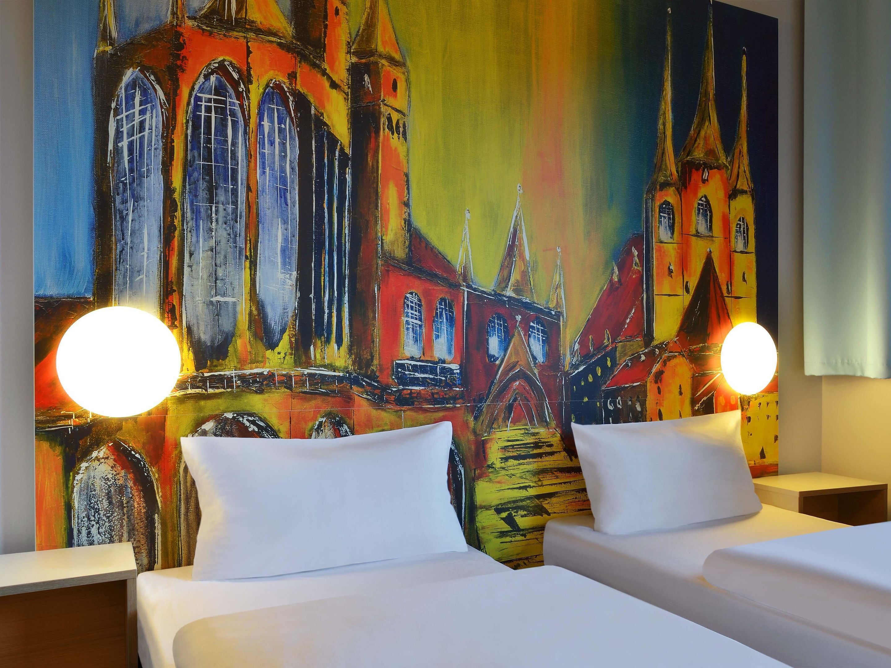 A hotel room with two beds, a colorful mural of a building, and two bedside lamps.