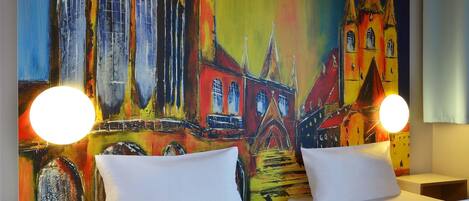 A hotel room with two beds, a colorful mural of a building, and two bedside lamps.
