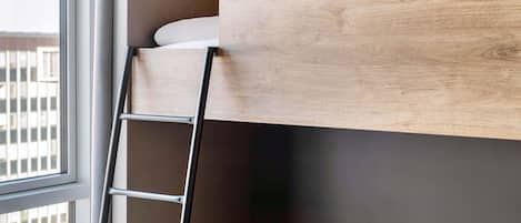 Een stapelbed met een houten hoofdbord, een metalen trap en een raam met uitzicht op gebouwen.