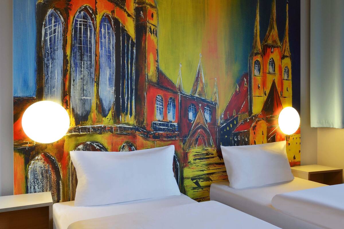 Une chambre d’hôtel avec deux lits, une fresque colorée représentant un bâtiment et deux lampes de chevet.