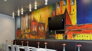 Un espace repas moderne avec des chaises et des tables blanches, une grande fresque colorée sur le mur et un téléviseur à écran plat fixé au-dessus des sièges.