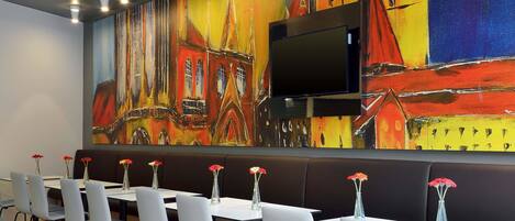 A modern dining area with white chairs and tables, a large colorful mural on the wall, and a flat-screen TV mounted above the seating.