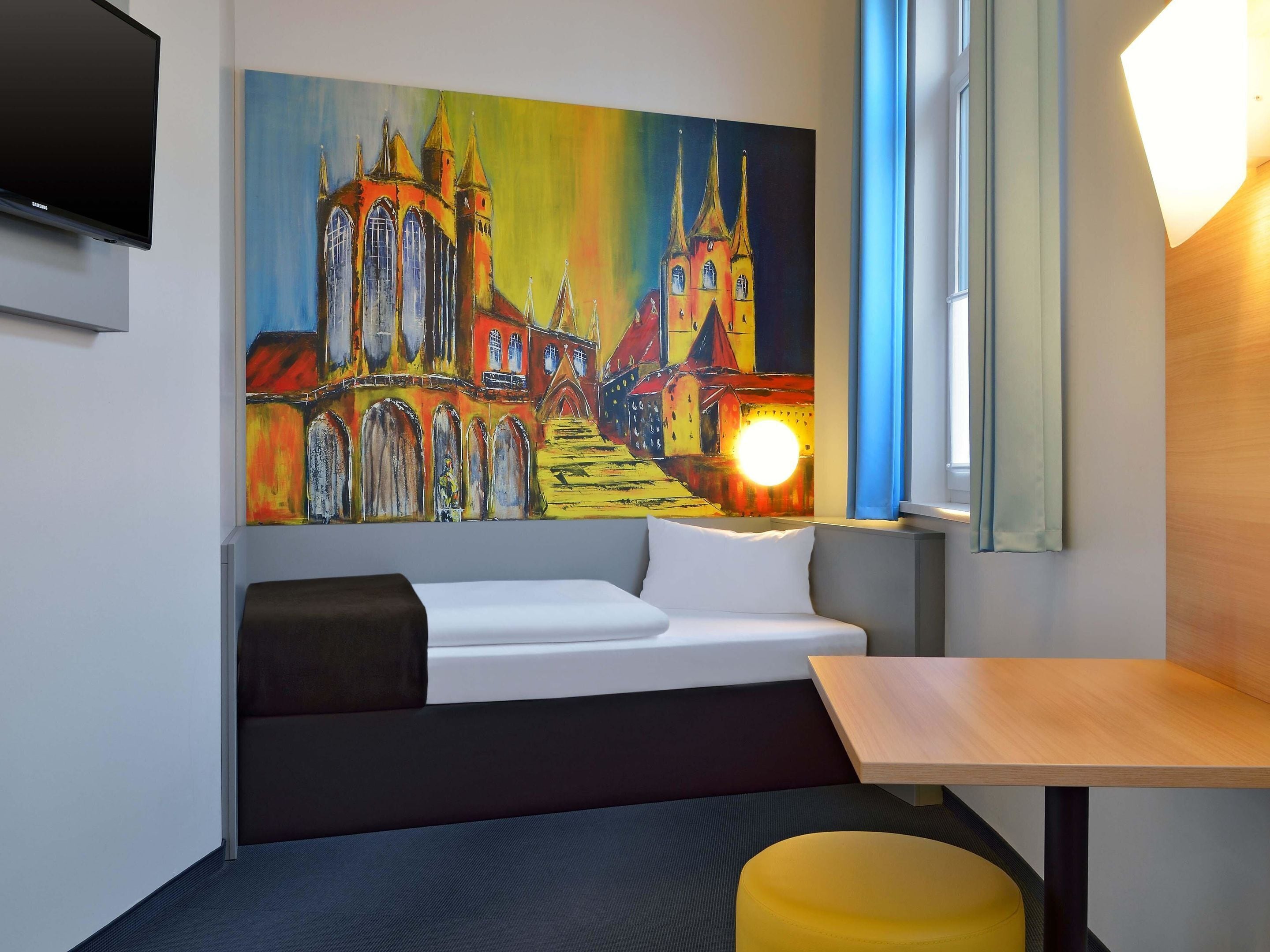 Una habitación de hotel moderna con un cuadro grande y colorido de un edificio, un sofá, una mesita y un televisor fijado a la pared.