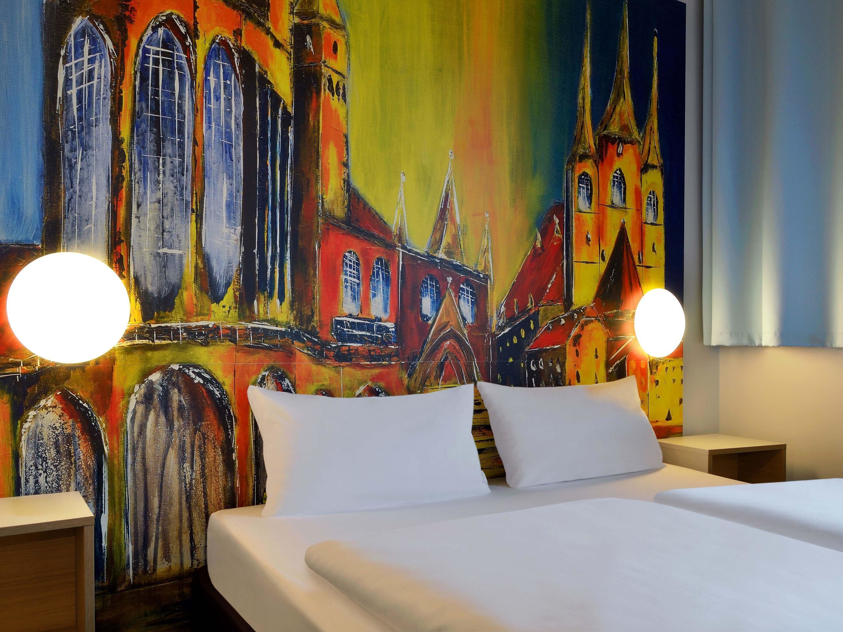 Una habitación de hotel con una cama grande, dos mesitas de noche y un mural colorido de un edificio en la pared.