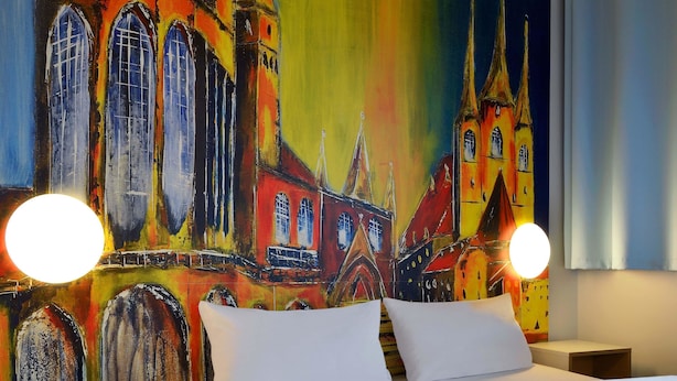 Une chambre d’hôtel avec un grand lit, deux tables de chevet et une fresque colorée représentant un bâtiment sur le mur.