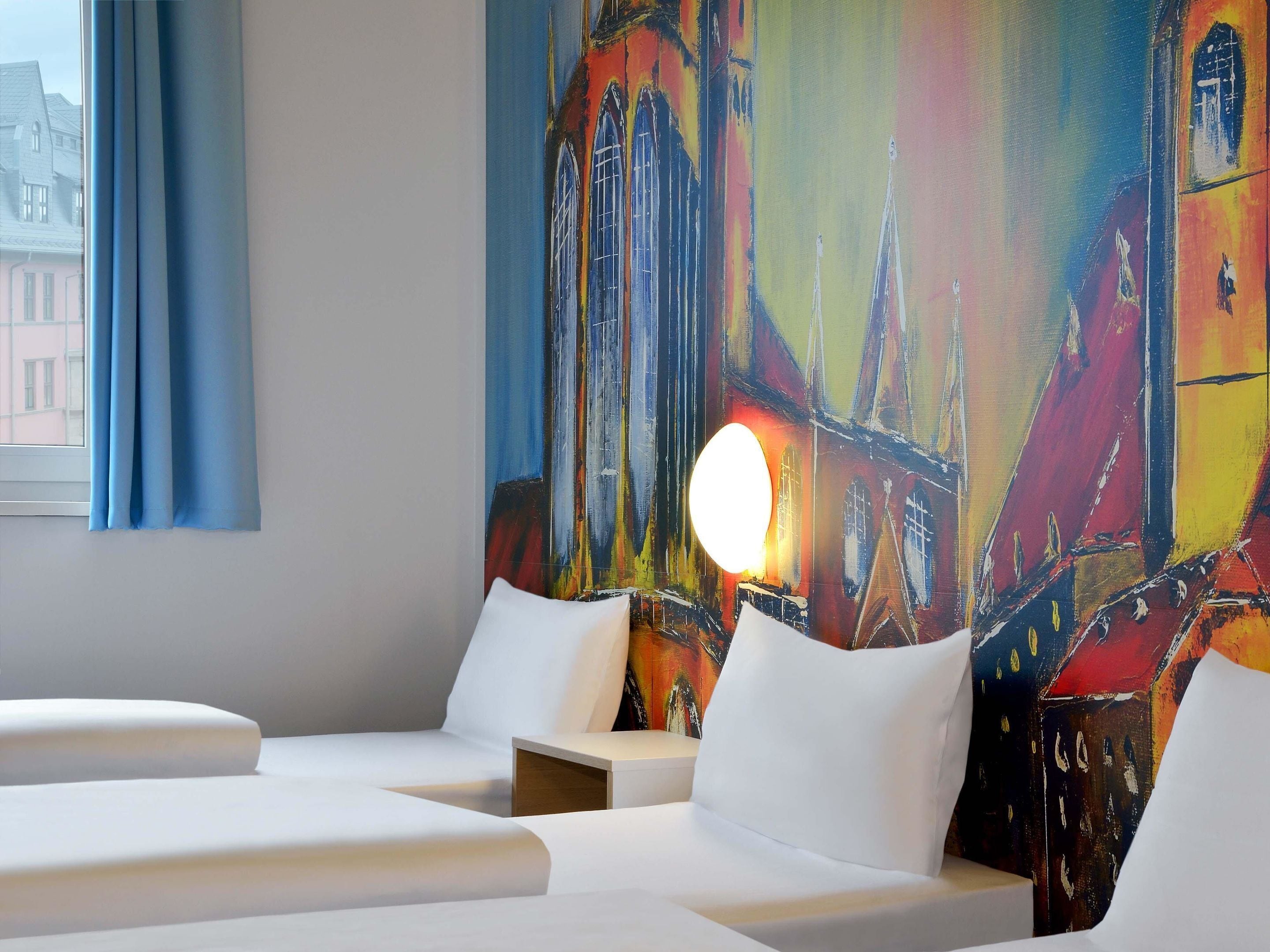 A hotel room with a large abstract painting on the wall, a window with blue curtains, and a bedside table with a lamp.