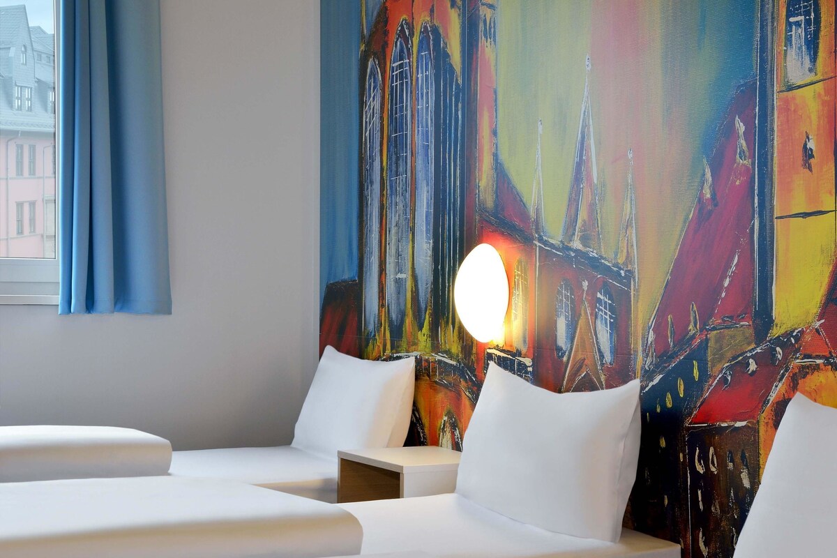 A hotel room with a large abstract painting on the wall, a window with blue curtains, and a bedside table with a lamp.