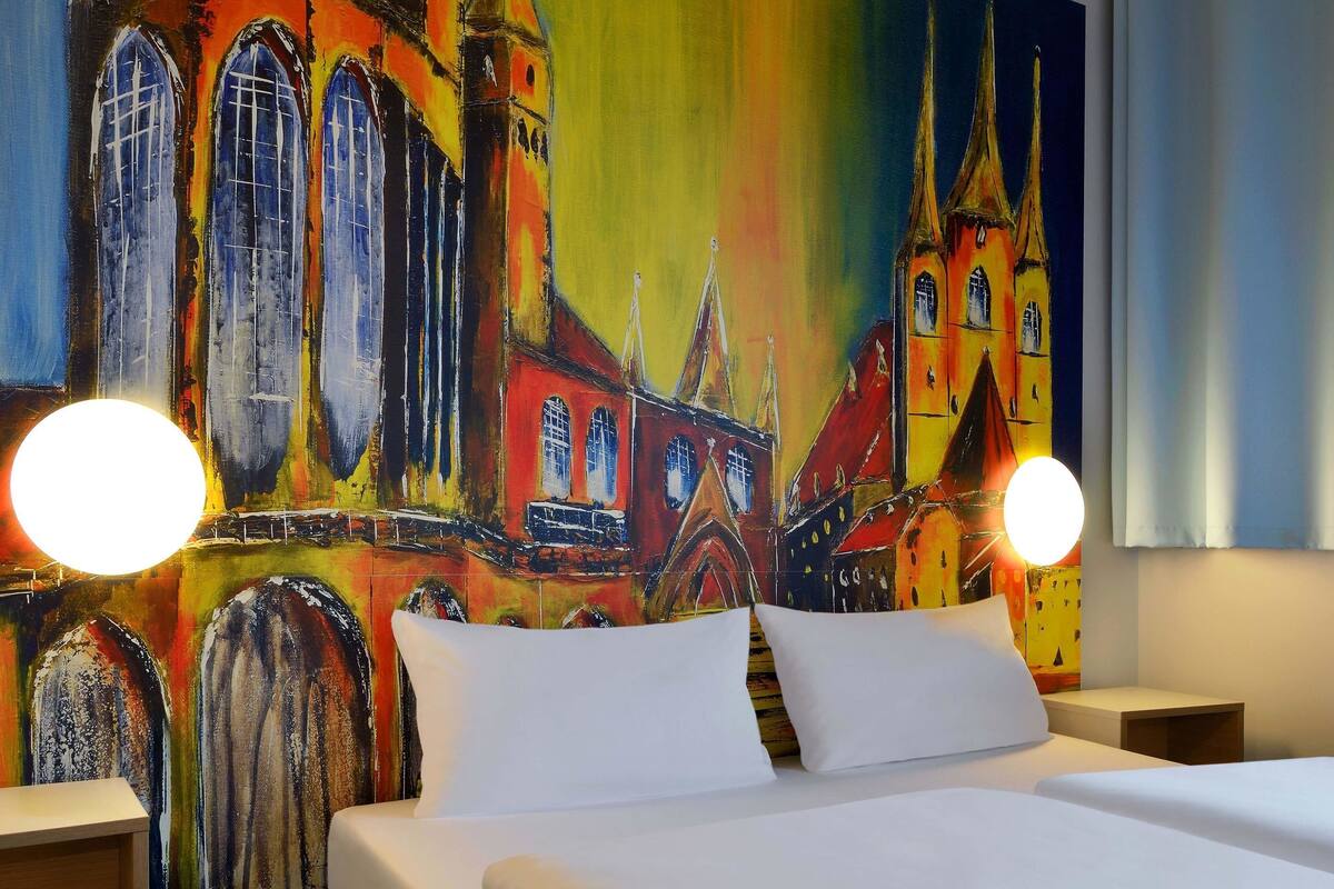 Une chambre d’hôtel avec un grand lit, deux tables de chevet et une fresque colorée représentant un bâtiment sur le mur.