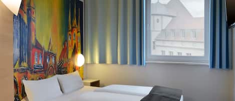 A hotel room with a bed, a desk, a painting of a building, and a window with curtains.