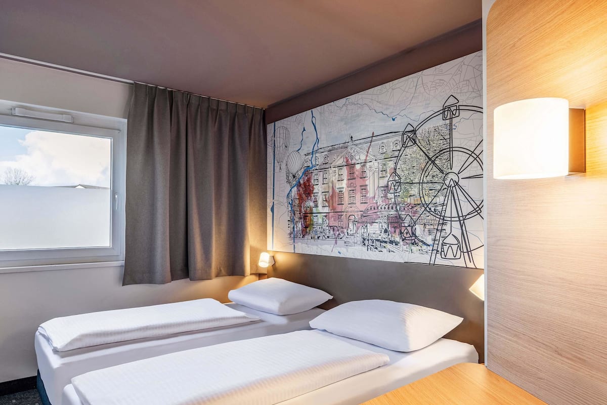A hotel room with two beds, a large map on the wall, a window with curtains, and a nightstand with a lamp.