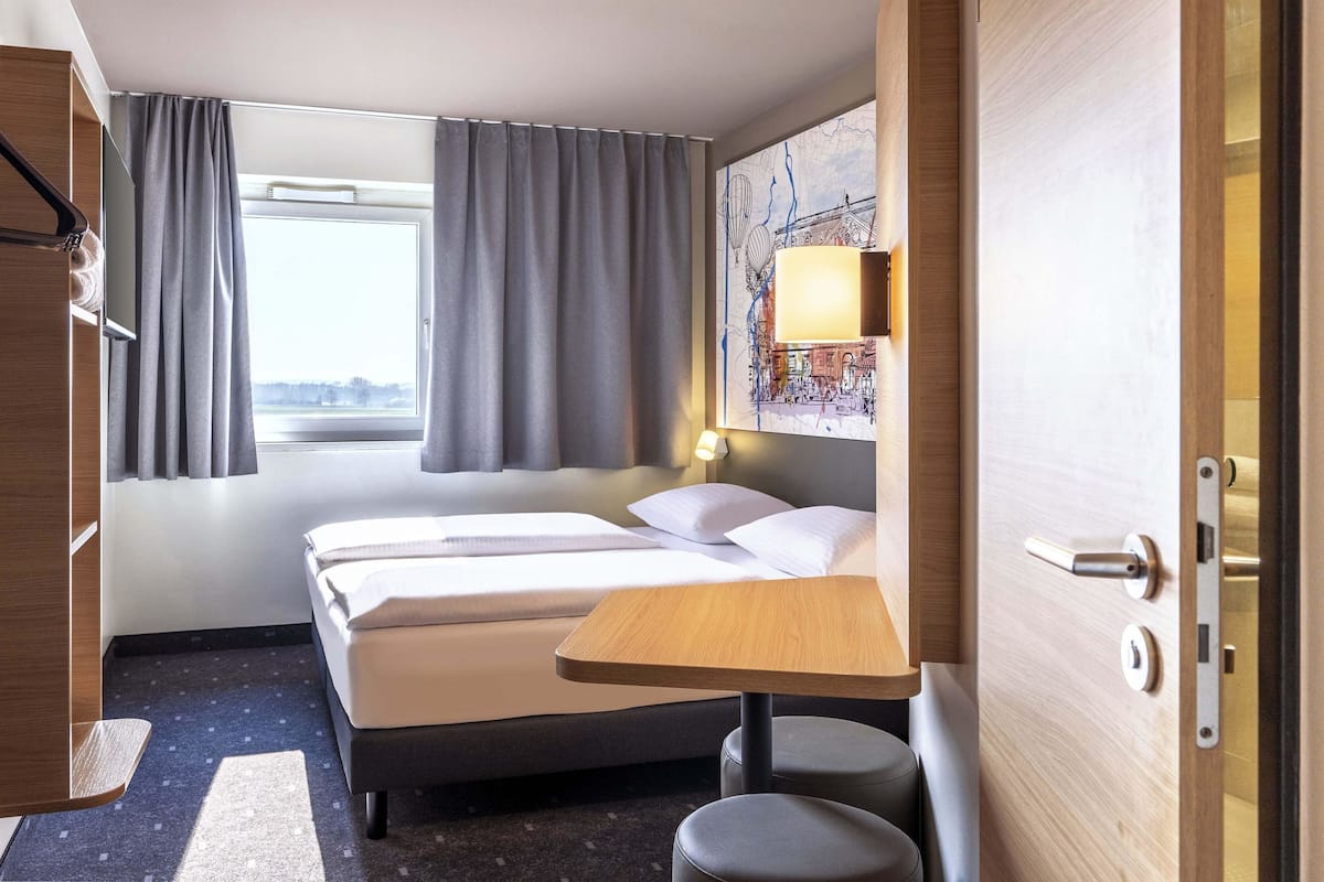A hotel room with a bed, a small table, a chair, and a window with curtains.