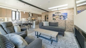 A modern living room with a grey sofa, a coffee table, and a patterned rug. There is a kitchen area with wooden cabinets and a dining set.