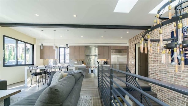 A modern kitchen with a stainless steel refrigerator, a dining area with a gray sofa and chairs, and a staircase with a metal railing leading to an upper level.