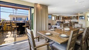 A spacious dining area with a large wooden table set for six, surrounded by upholstered chairs. A kitchen area is visible in the background, and the room has large windows offering a view of the outside.