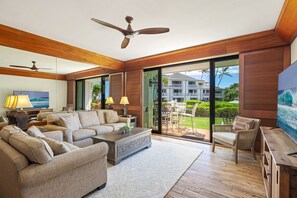 A spacious living room with a sofa set, a coffee table, and a TV. There is a ceiling fan, a sliding glass door leading to a balcony, and a view of a building with a pool. - Poipu Kapili 16: Ocean Views; Fully Updated-New Furnishings, Split ACs & Windows (Koloa)