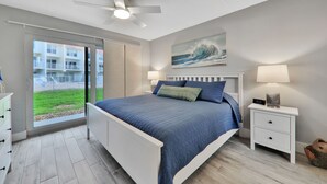 Een slaapkamer met een bed, nachtkastjes, een plafondventilator, een raam met uitzicht op een gebouw en een schilderij aan de muur.