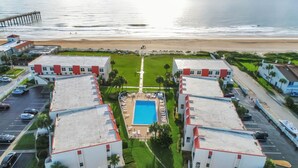 Een hotel met een centraal gelegen zwembad, omringd door parkeerplaatsen en uitzicht op het strand.