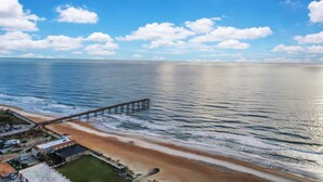 Een strand met een pier die in de oceaan reikt, een parkeerplaats en een gebouw met een zwembad.