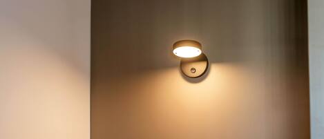 A neatly made bed with a headboard against a plain wall, a wall-mounted light fixture, and a small circular object on the wall.