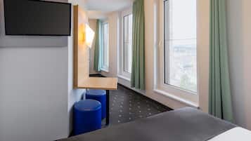 Habitación de hotel con cama, televisor montado en la pared, pequeño escritorio y ventanas grandes con cortinas.