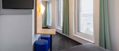 Habitación de hotel con cama, televisor montado en la pared, pequeño escritorio y ventanas grandes con cortinas.