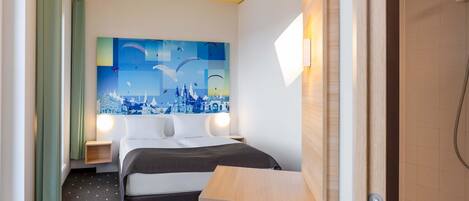 A hotel room with a bed, a nightstand, a wall-mounted light, a wooden table, and a blue trash bin.