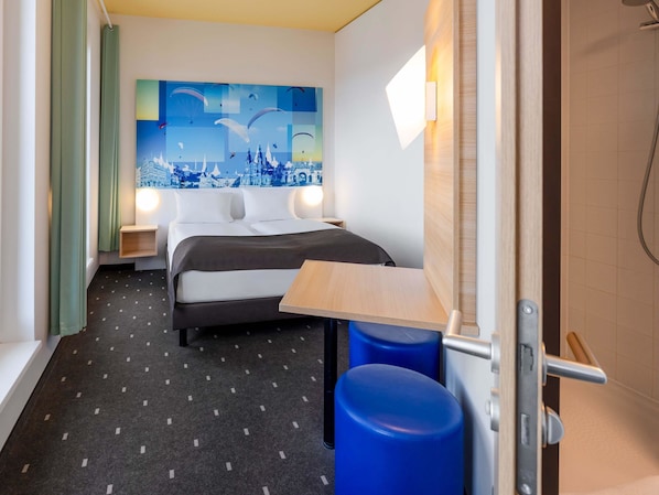 A hotel room with a bed, a nightstand, a wall-mounted light, a wooden table, and a blue trash bin. - B&B HOTEL Fulda-City (Fulda)