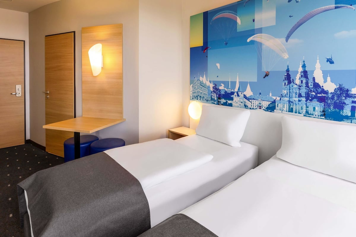 A hotel room with two beds, a wooden desk, a wall-mounted light, and a cityscape mural.