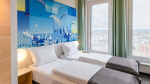 Habitación de hotel con dos camas, un ventanal con vistas a la ciudad y un mural en la pared que representa un paisaje urbano.