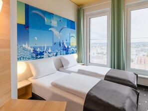 A hotel room with two beds, a large window with a view of the city, and a wall mural of a cityscape. - B&B HOTEL Fulda-City (Fulda)
