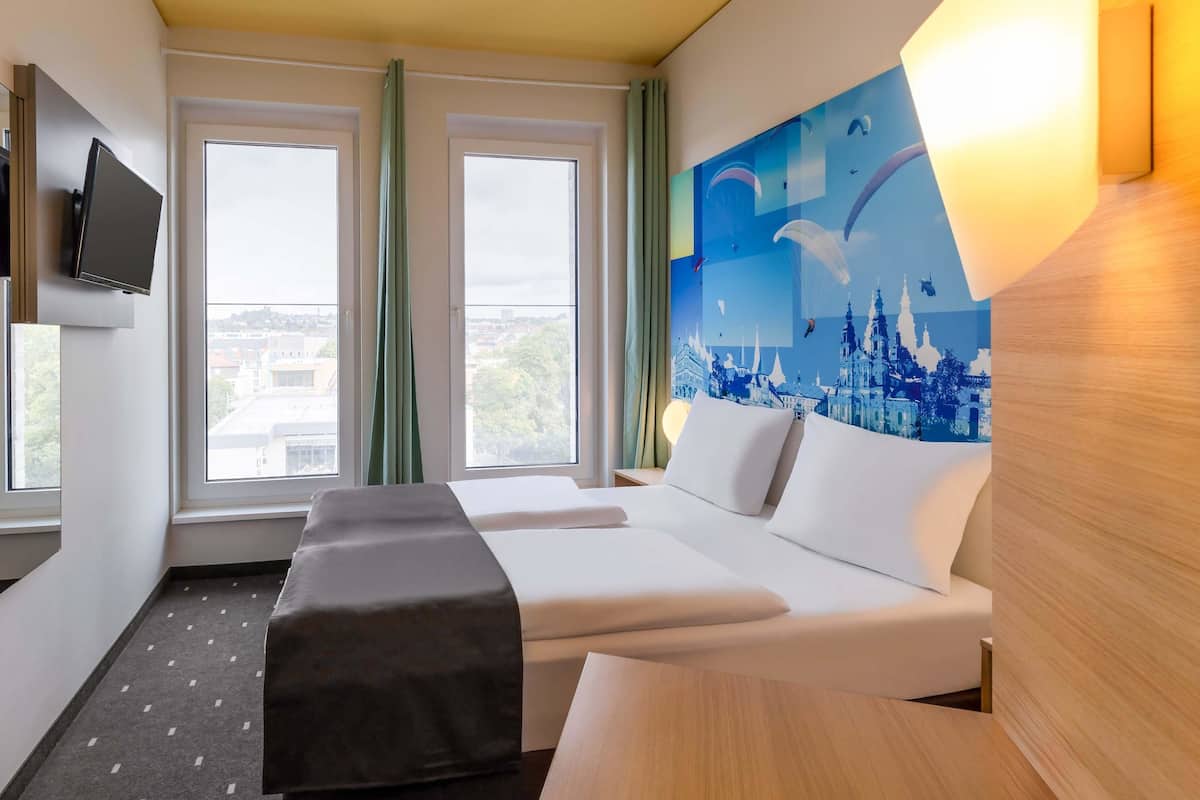 A hotel room with a bed, a desk, a TV, and a painting of a cityscape.