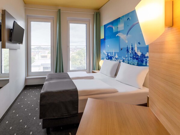 A hotel room with a bed, a desk, a TV, and a painting of a cityscape. - B&B HOTEL Fulda-City (Fulda)