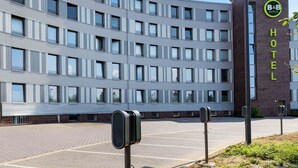 Ein modernes Hotelgebäude mit einem gut sichtbaren 'B&B Hotels'-Schild, einem Parkplatz und einer Sicherheitswache.