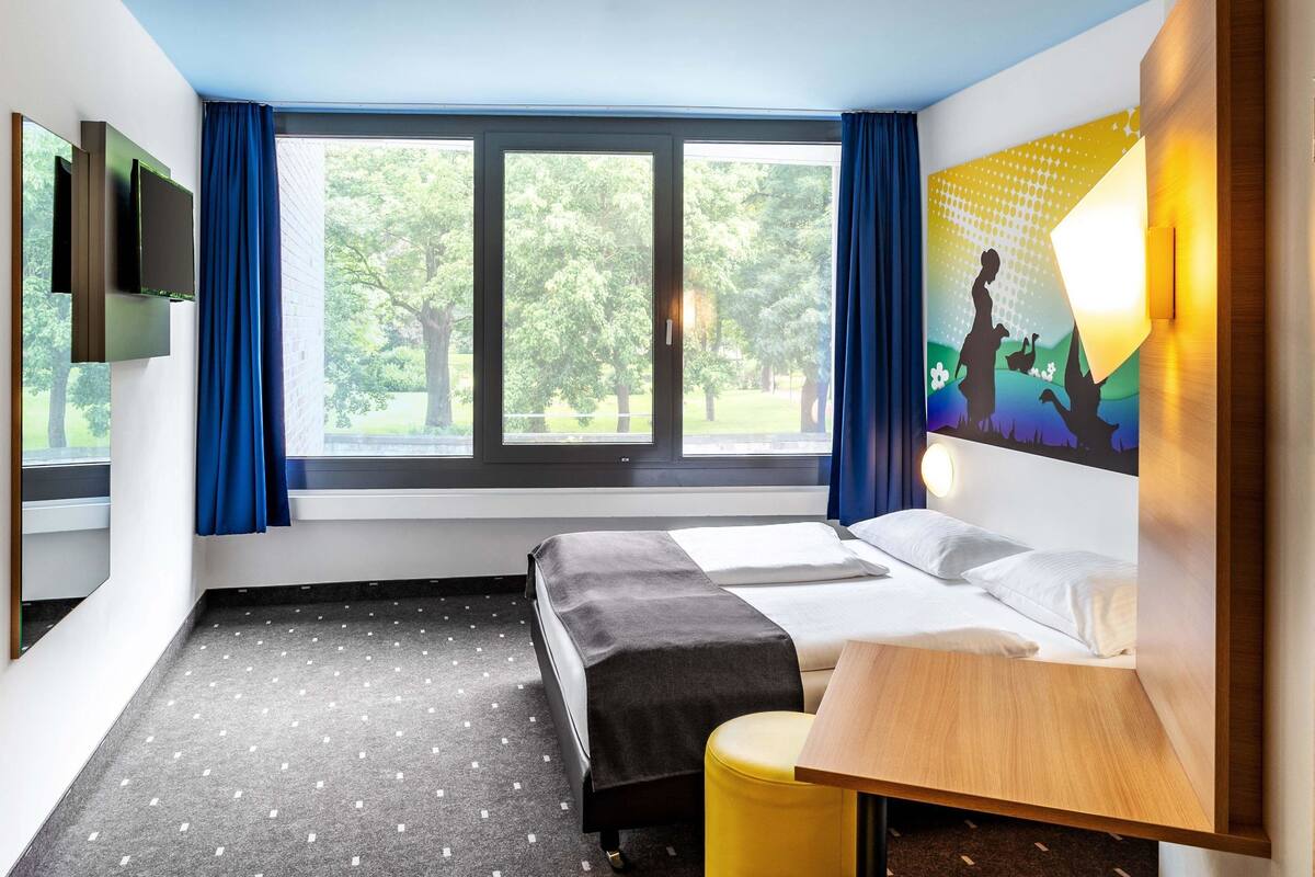 Ein Hotelzimmer mit einem Bett, einem kleinen Tisch, einem Stuhl, einem Fernseher und einem Fenster mit Vorhängen.