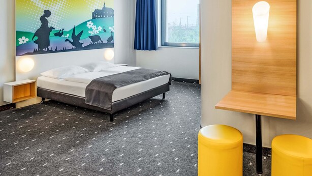 Une chambre d’hôtel avec un lit, une lampe fixée au mur, un tableau et une fenêtre avec des rideaux.