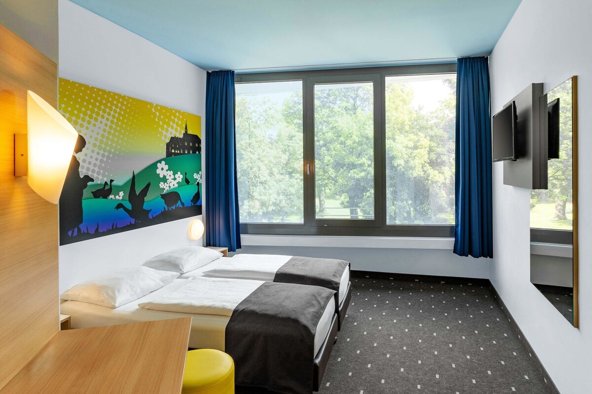Une chambre d’hôtel avec deux lits, un bureau, une chaise, une télévision et une grande fenêtre avec des rideaux.