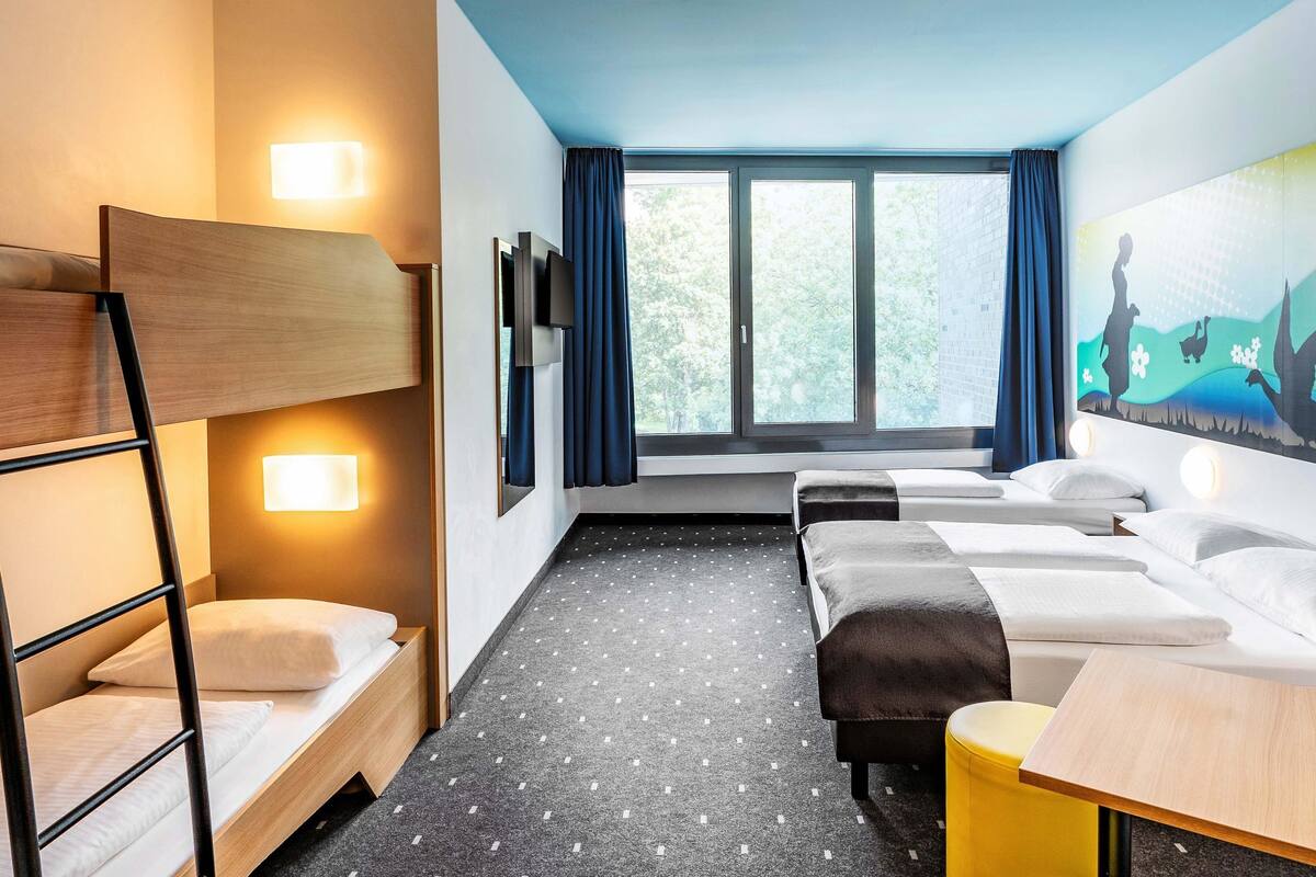 Ein Hotelzimmer mit Etagenbett, zwei Einzelbetten, einem Schreibtisch und einem großen Fenster mit Vorhängen.