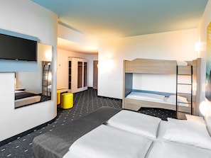 A hotel room with a bunk bed, a flat-screen TV mounted on the wall, a small table with a chair, and a yellow luggage trolley.