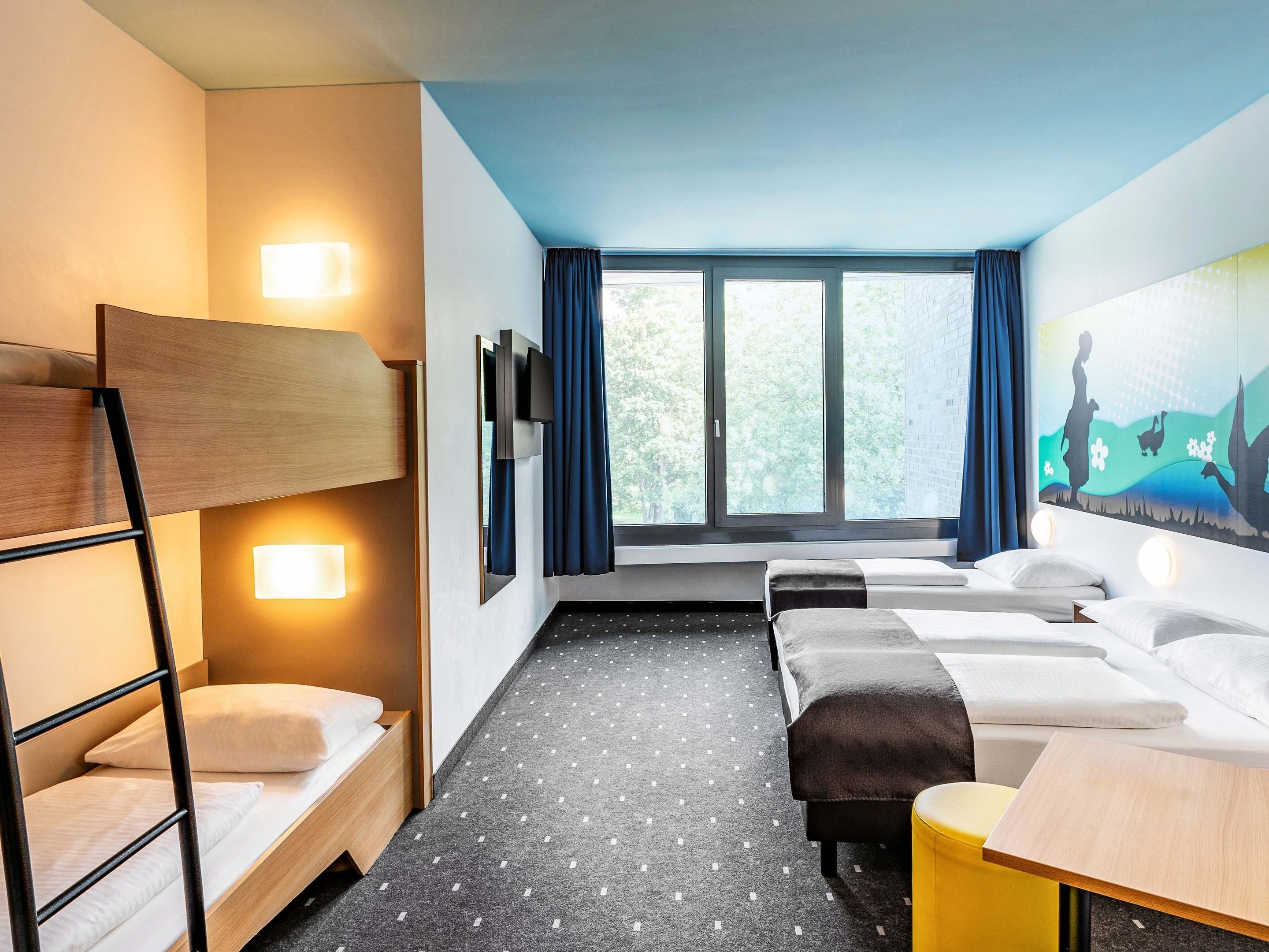 Ein Hotelzimmer mit Etagenbett, zwei Einzelbetten, einem Schreibtisch und einem großen Fenster mit Vorhängen.