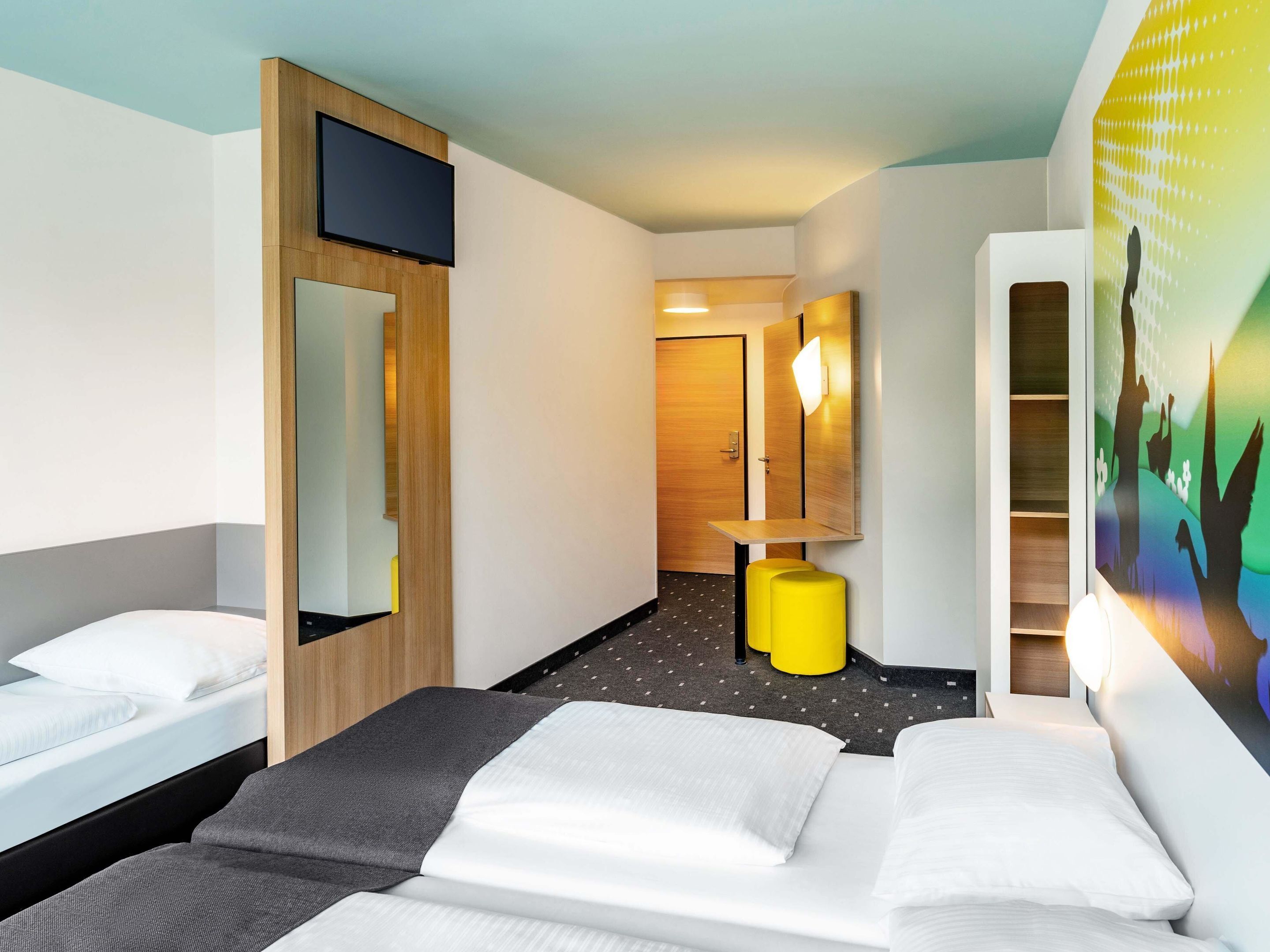 Ein Hotelzimmer mit Bett, Schreibtisch, Spiegel und einem am Wand montierten Fernseher.