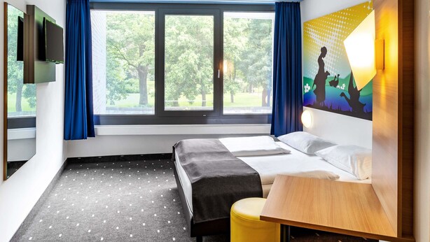 Ein Hotelzimmer mit einem Bett, einem kleinen Tisch, einem Stuhl, einem Fernseher und einem Fenster mit Vorhängen.
