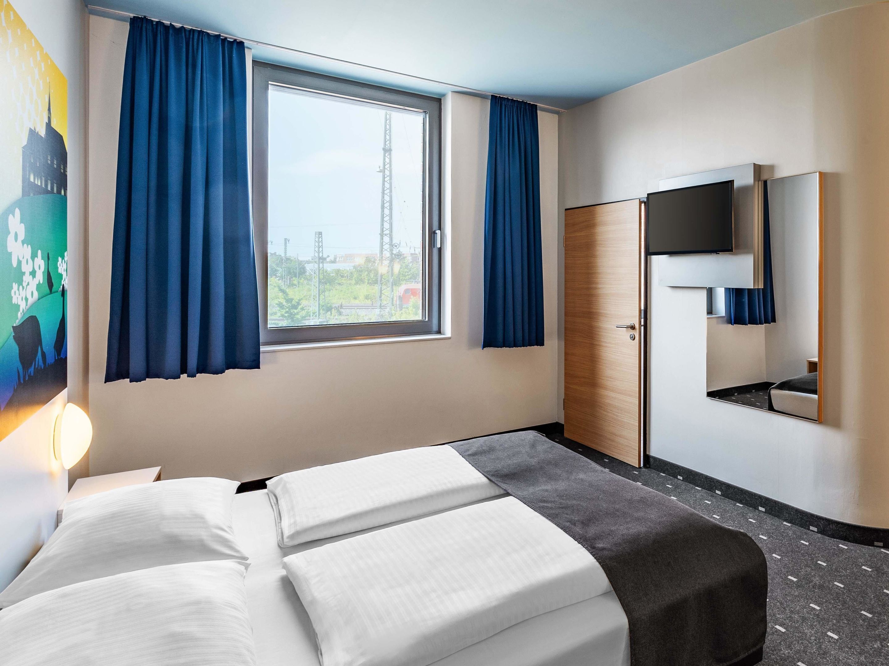 Una habitación de hotel con cama, televisor, armario y ventana con cortinas.