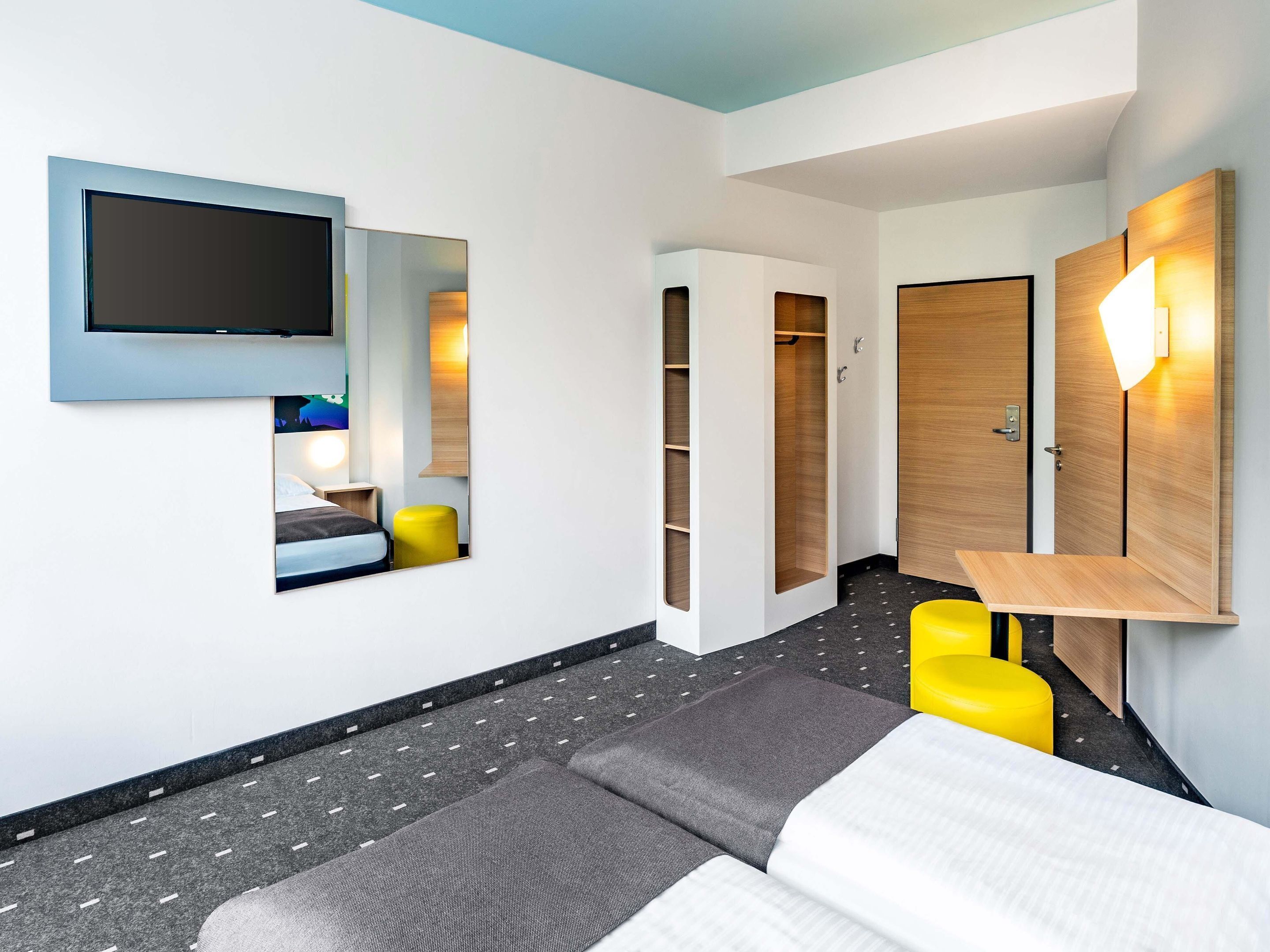 Una habitación de hotel moderna con una cama, un televisor montado en la pared, un armario y una pequeña mesa.