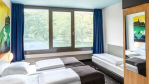 Ein Hotelzimmer mit zwei Betten, einem großen Fenster mit Vorhängen, einem Spiegel und Blick auf Grünfläche.