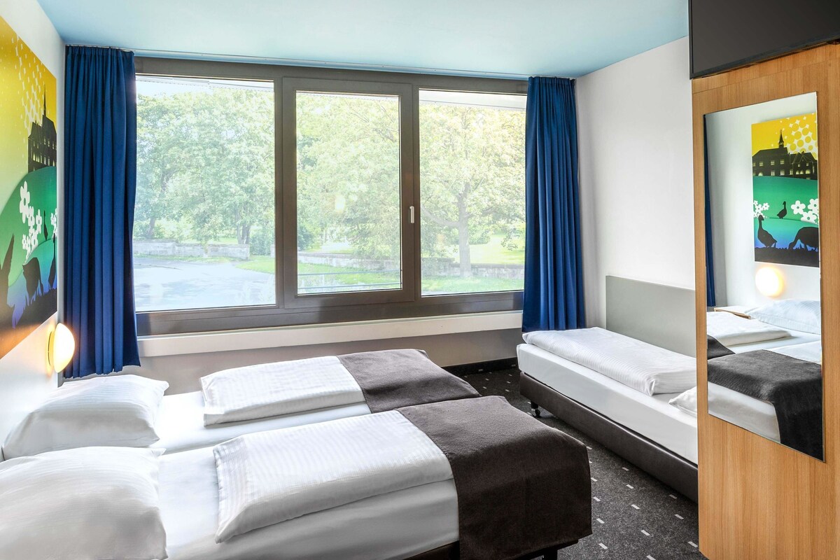 Une chambre d’hôtel avec deux lits, une grande fenêtre avec des rideaux, un miroir et une vue sur la verdure.