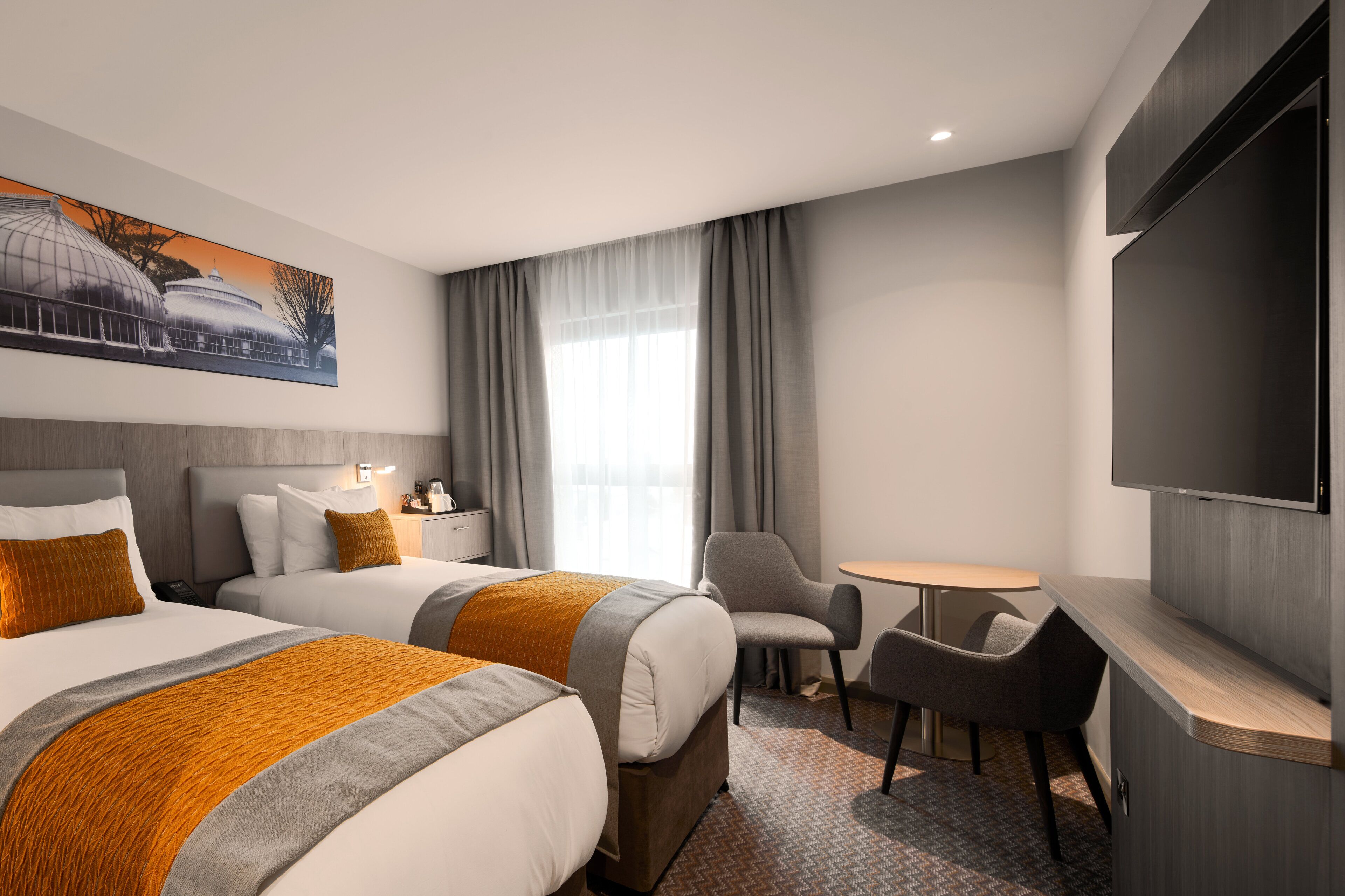 A hotel room with two beds, a desk, a chair, a TV, and a large window with curtains.