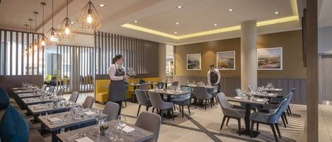 Un restaurante moderno con varias mesas de comedor, algunas con sillas y otras con asientos acolchados amarillos. Hay personal de servicio presente, y la sala tiene un diseño contemporáneo con iluminación colgante y obras de arte en las paredes.